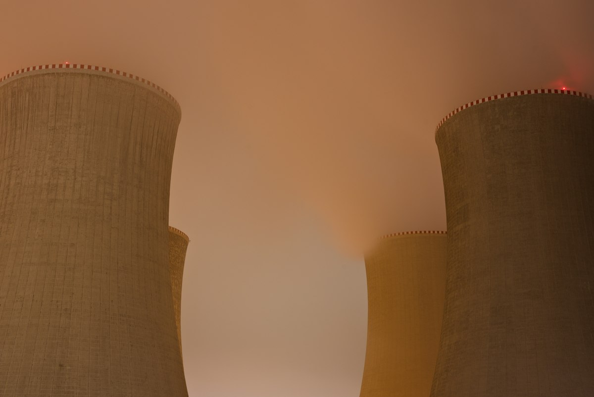 Hyperbolic natural-draft evaporative cooling towers at a thermal power station at night, illuminated against an atmospheric foggy sky, visualising the industrial scale of heat rejection these systems handle. Photo: Lukáš Lehotský / Unsplash